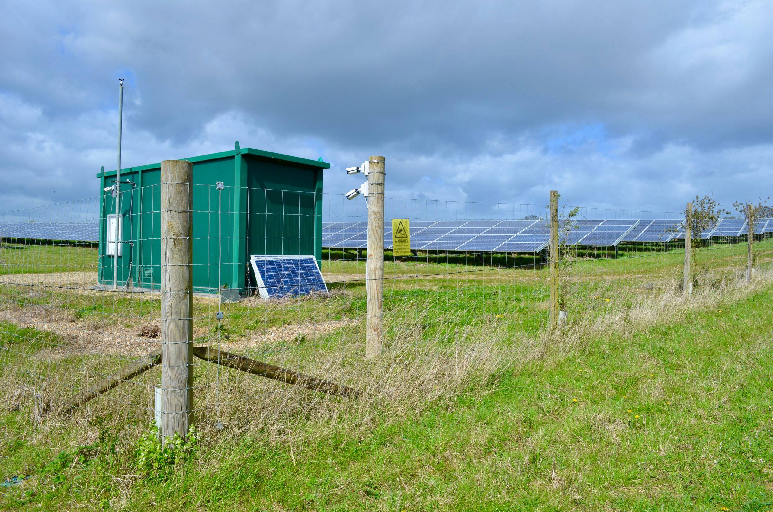 A rural solar farm with a green equipment shed and solar panels under a cloudy sky.